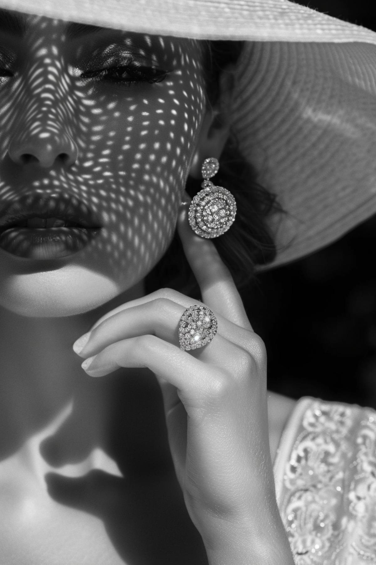 A close-up of the hand wearing an elegant diamond ring and earrings, with her face partially obscured by a white hat, showcasing luxury jewelry in monochrome tones.