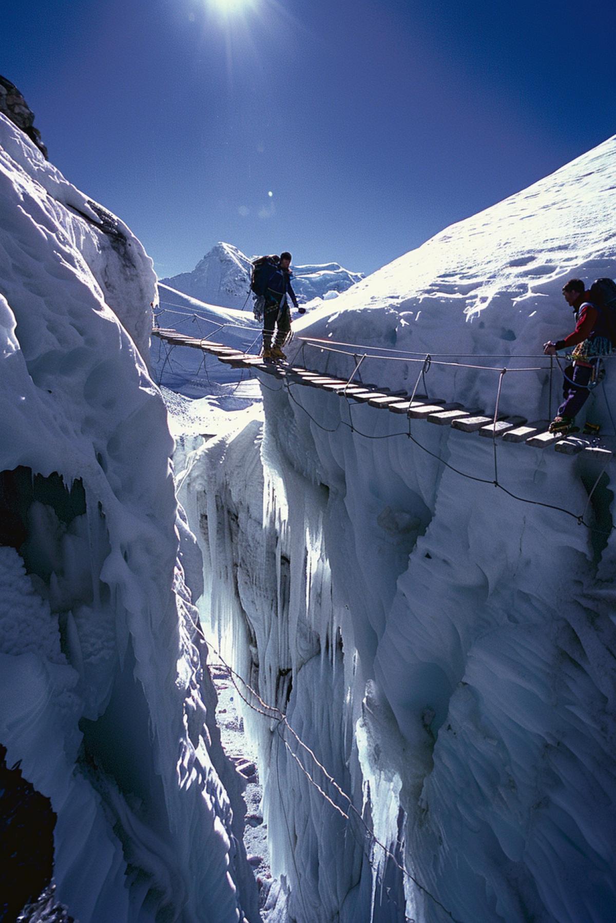 Climbers on a bridge over an ice crack, deep ice canyon sparkling in the sun, wooden bridge, ropes, climbers with heavy backpacks, blue sky, ice crack walls, deep shadows, foreground bridge and climbers, middle ground crack, background snowy peaks, tension and caution, the grandeur of nature, a sense of height and danger, courage and trust.