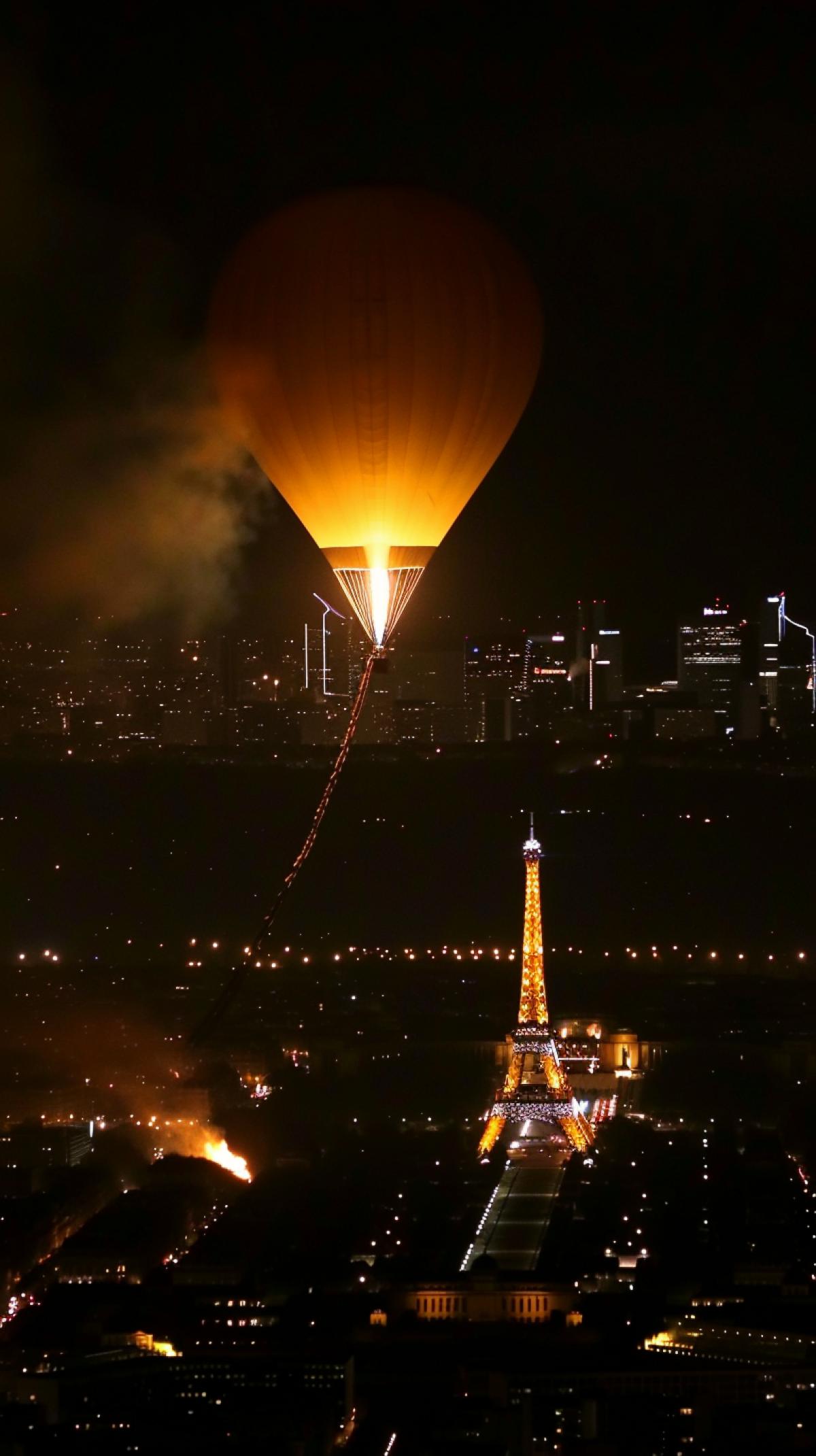 Olympic Games in Paris: night, birds eye view, the Eiffel Tower illuminated by spotlights, lanterns sparkling on it, a hot air balloon hovers in the sky nearby, along the rim of which the Olympic flame is burning, the outlines of the night city are unclear below