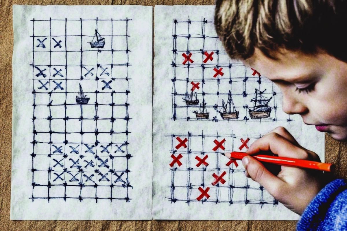 Children play a game on paper: sea battle, a boy holds a sheet of paper and a pencil, crosses are drawn on a sheet of paper to indicate ships, a sheet of paper is checkered