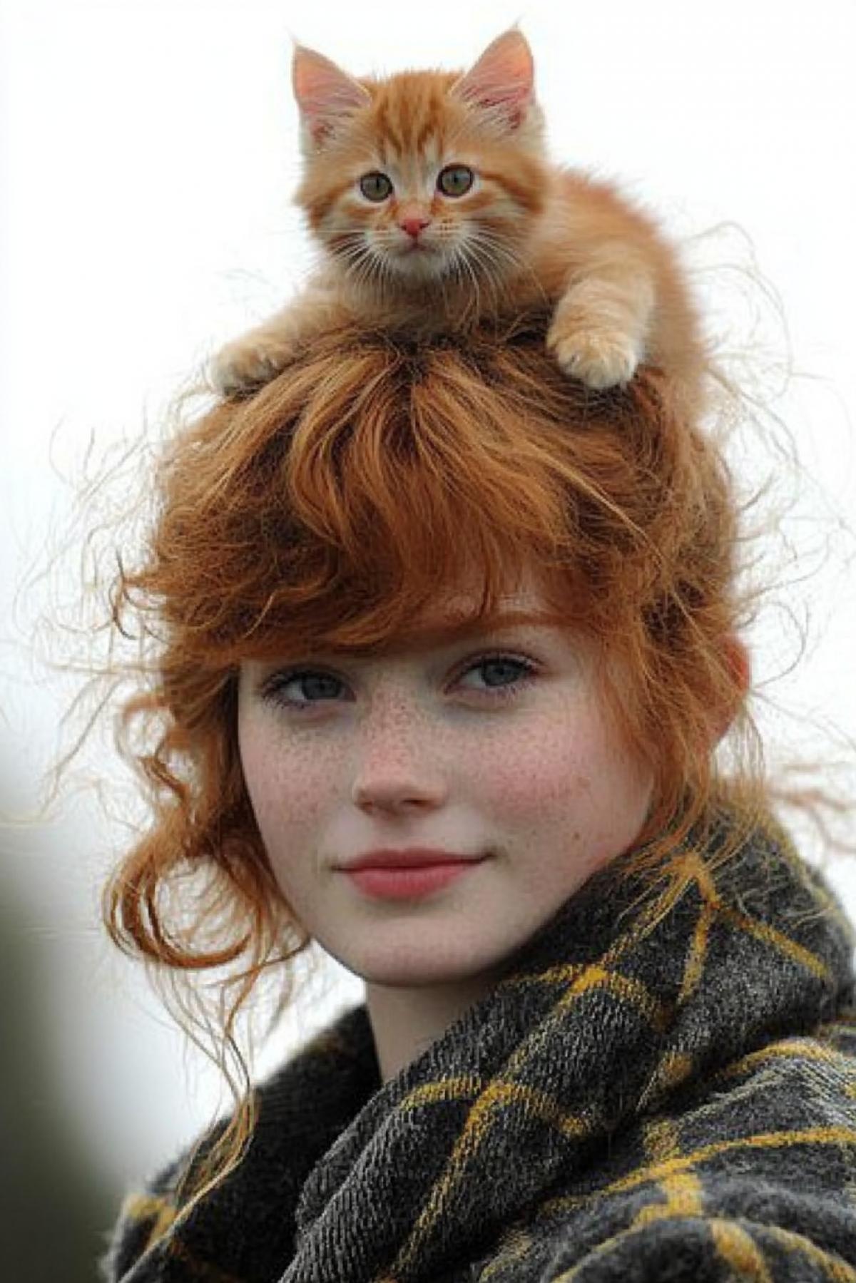 A shaggy redheaded girl. There is a red kitten hiding on her head, on the top of her head, in her hair.