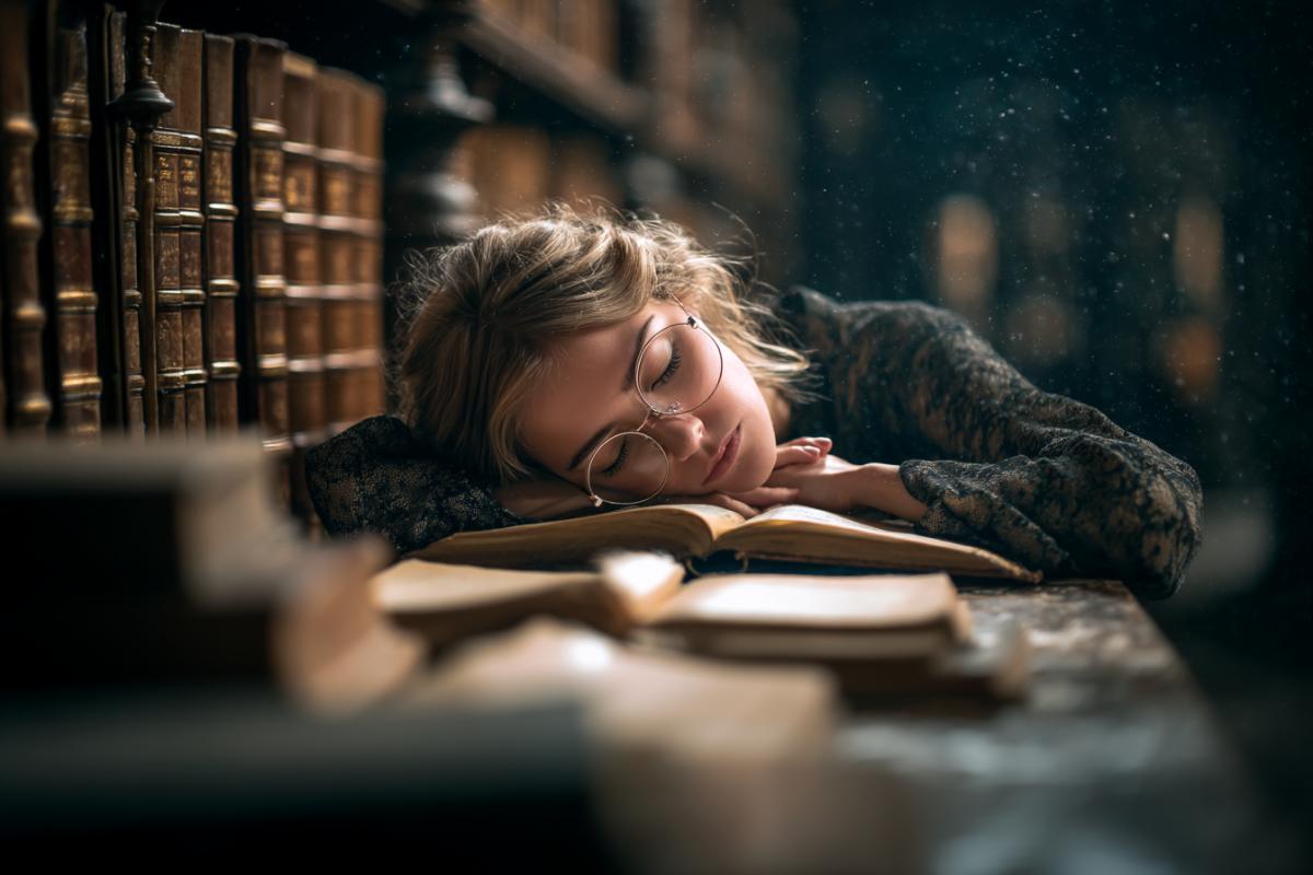 Ancient library. Between the rows of high book shelves a girl with glasses that fell asleep behind an open book. Her hand still holds the page. Light dust in the air.