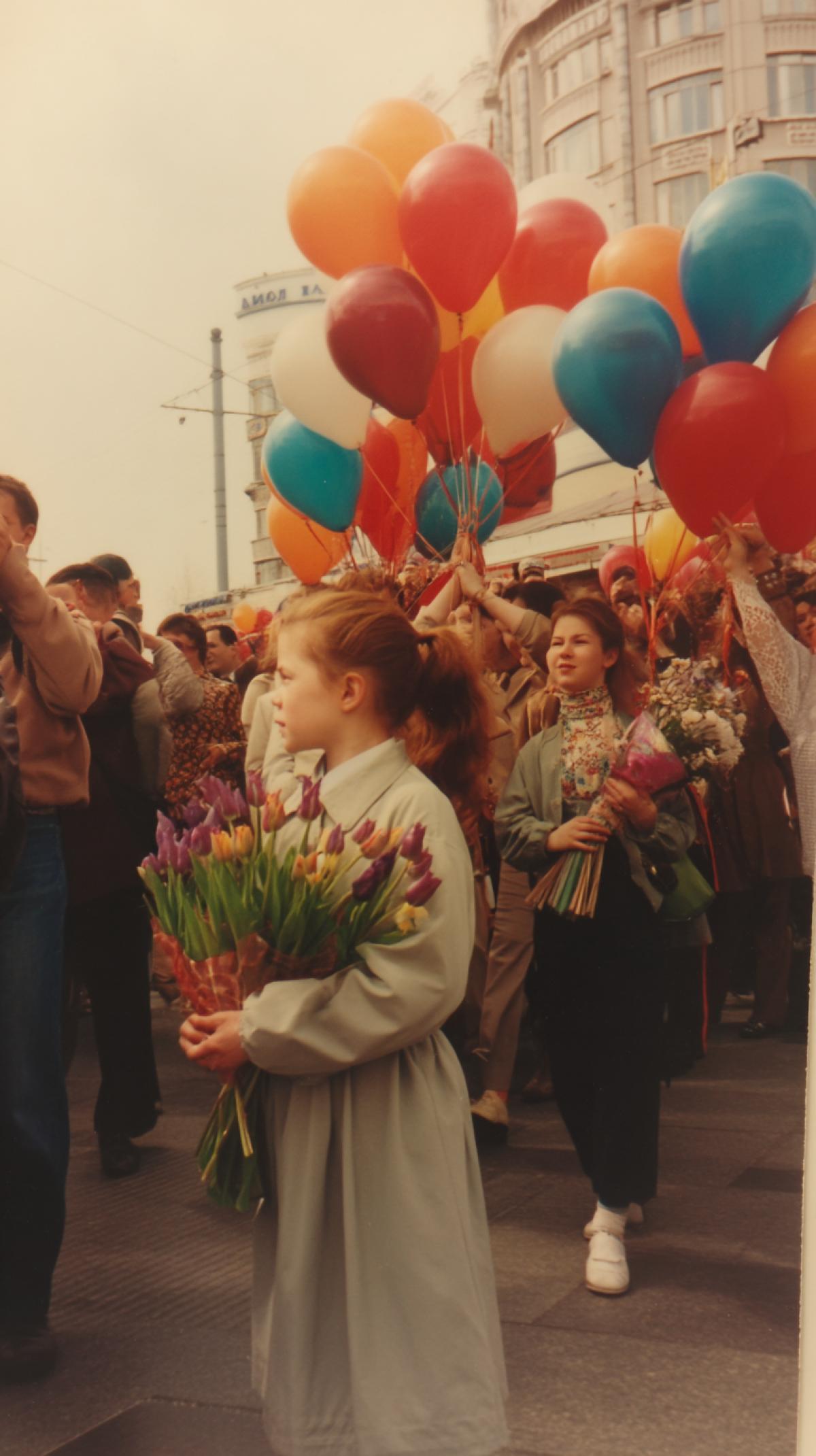 Photo of the 90s Russia. Victory Parade. Joyful people with balloons and bouquets of lilac and tulips. Girl 5 years old in the foreground. The photo is taken from the lower angle.