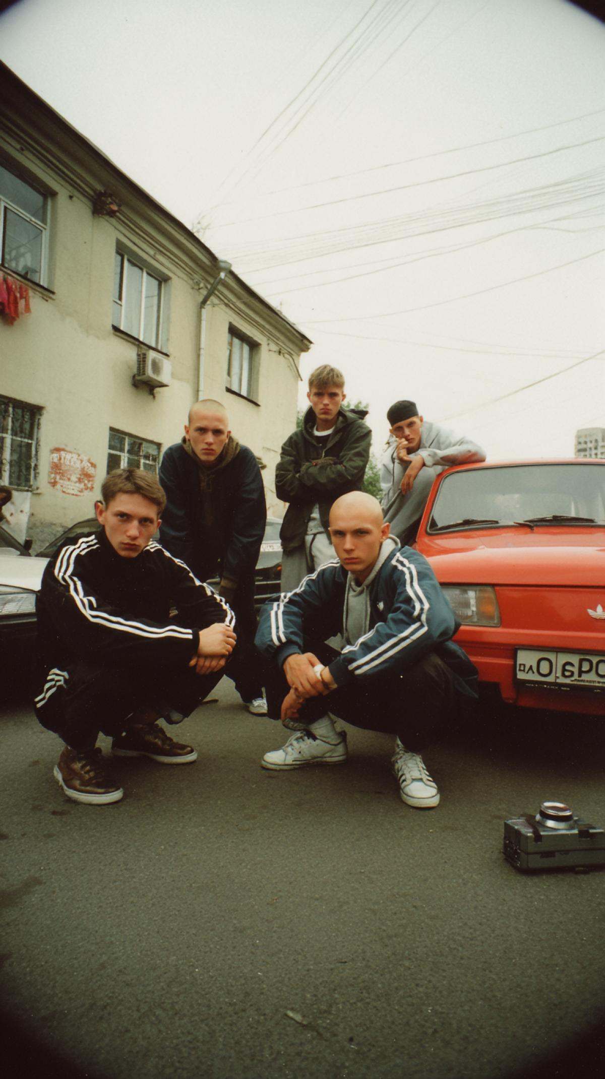Photo of the 90s Russia. A group of impudent young guys. Bald. They squat. In the costumes of Adidas and dark shoes with a sharp toe. Smoke. In front of them is a gray tape recorder. And behind the red car Lada Zhiguli