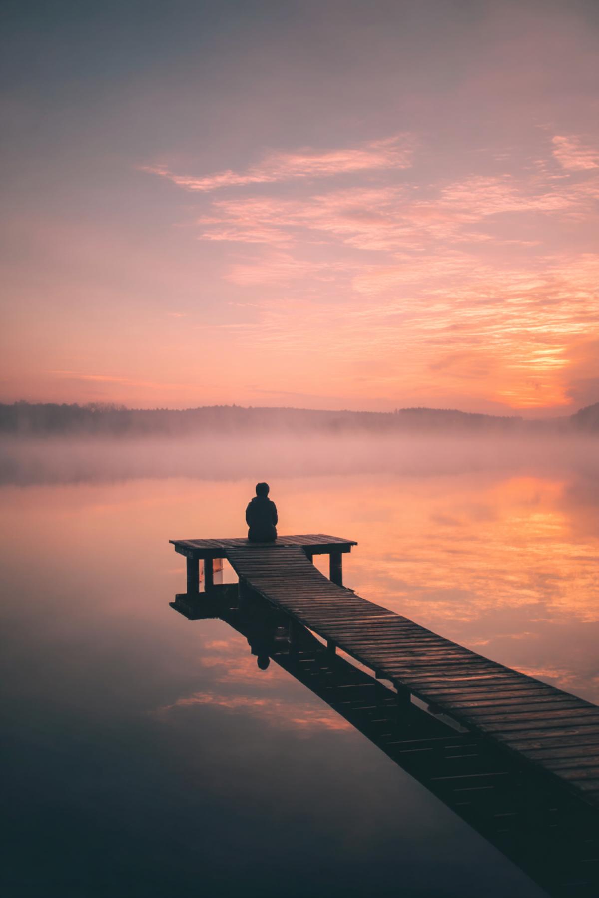 solitary figure sitting on a wooden dock, calm lake reflecting morning mist, pastel sunrise sky, soft ripples in the water, peaceful expression, absolute serenity