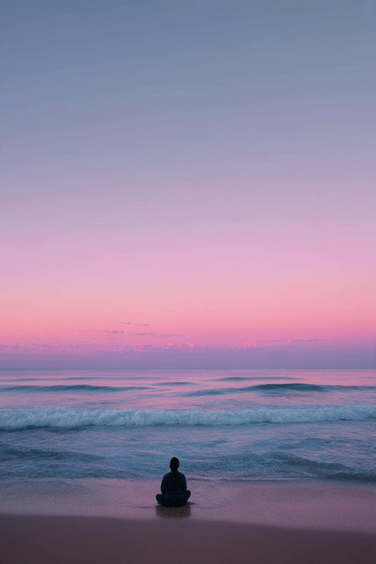 person sitting cross-legged on beach at dawn, watching soft waves roll in, wide horizon, pastel sky with subtle pink and blue, tranquil and meditative atmosphere