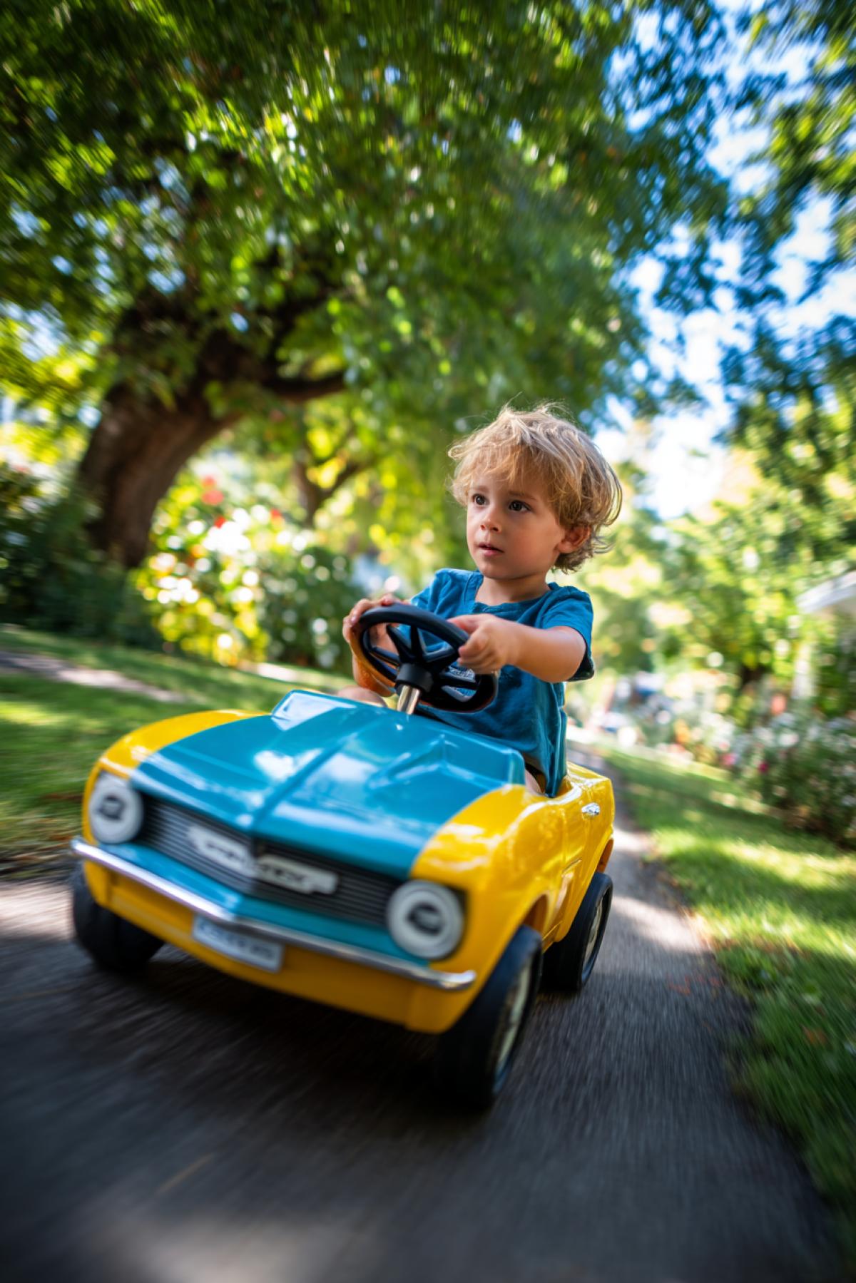 playful happy 4yo boy within small pedal car fashioned as Chevrolet comaro studying to drive 1970s city park environment happiness and carelessness bright summer sunny day colours trees flowers butterflies low view happy eyes in focus high contrast
