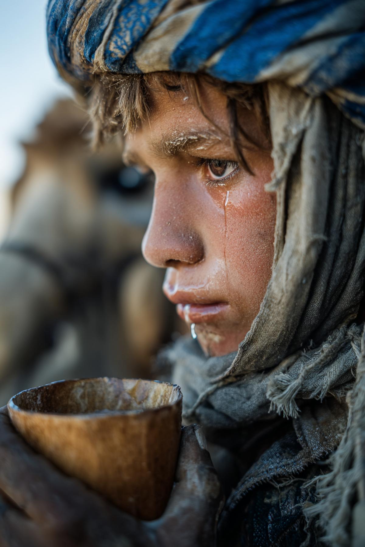 Close-up of a young boy drinking from a wooden cup in the caravan, his lips cracked from the salt wind, a drop of water running down his chin. Over his shoulder a blurred figure adjusting a load on a camel. Dusty palette with a single vivid touch: the blue stripe on his faded headscarf. Documentary style, raw and un4kd