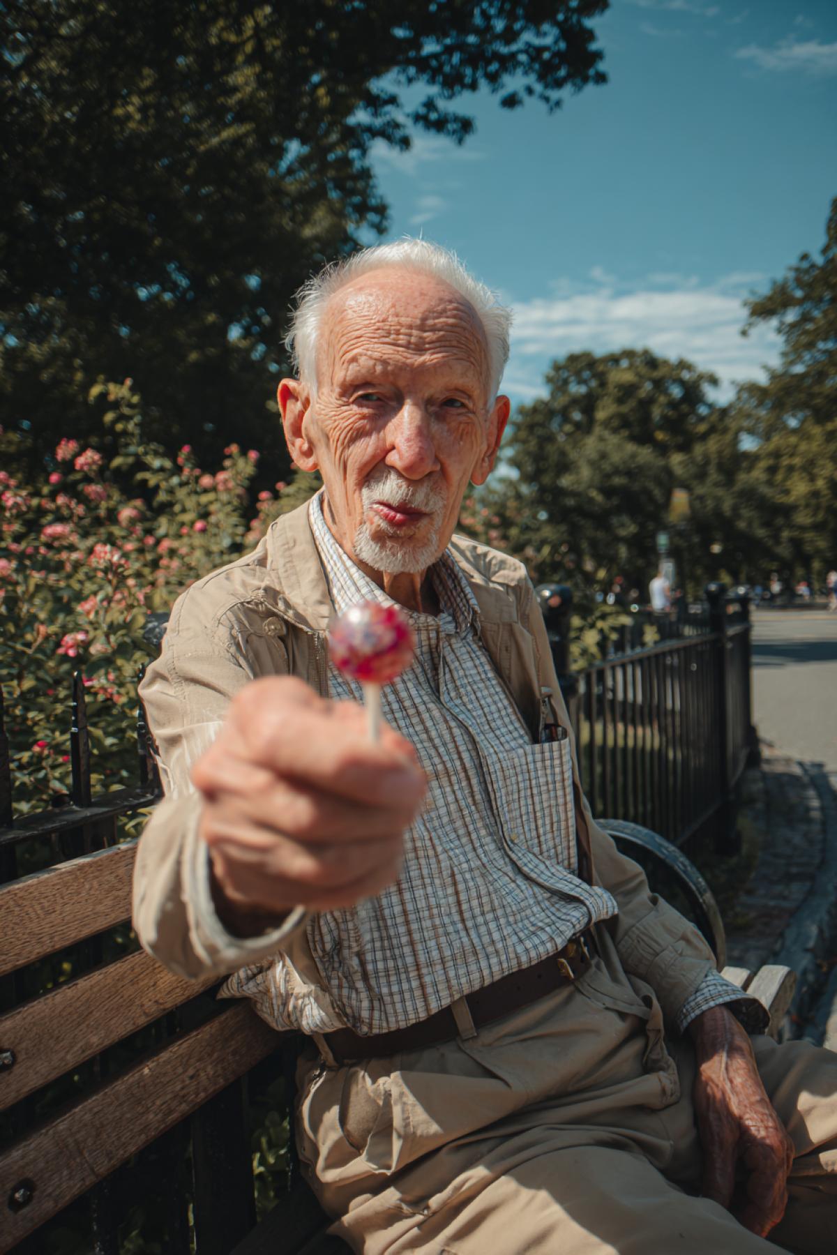 A sunny morning in a city park. A neat, very kind-looking elderly man is sitting on a bench, holding out a beautiful and delicious candy directly toward the camera.