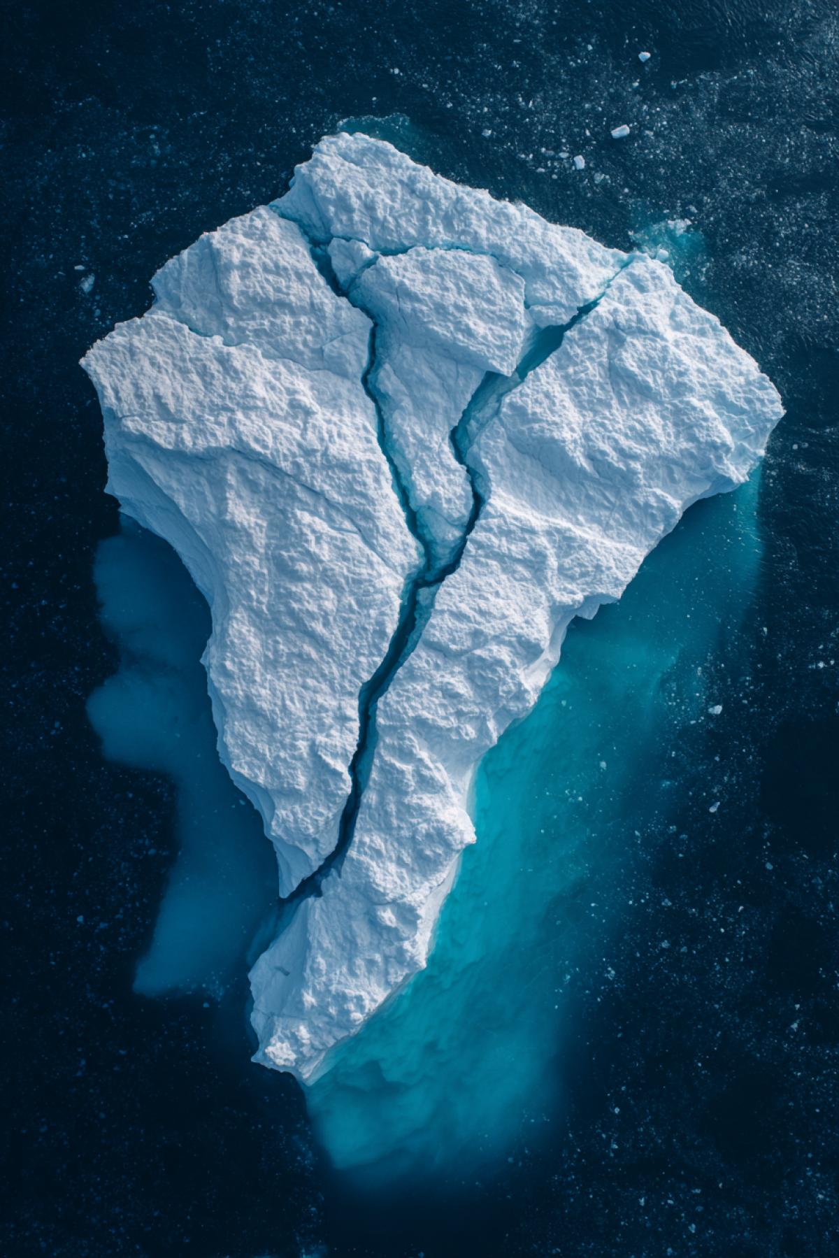Top-down photo of a big iceberg in Arctic Ocean, 90% of its mass visible underwater, turquoise cracks contrasting with dark blue sea, shot from helicopter, Nikon Z9, climate change symbolism, chilling beauty.