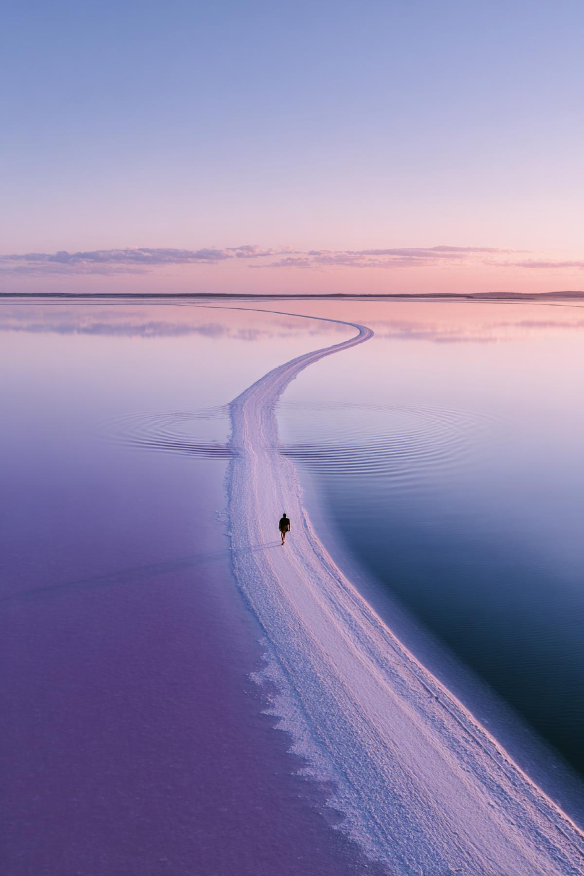 Abstract top-down shot of pink salt flats in Australia, shallow water creating perfect reflections, lone walker in center leaving ripple trails, minimalistic com4k, Hasselblad X2D, surreal pastel tones.