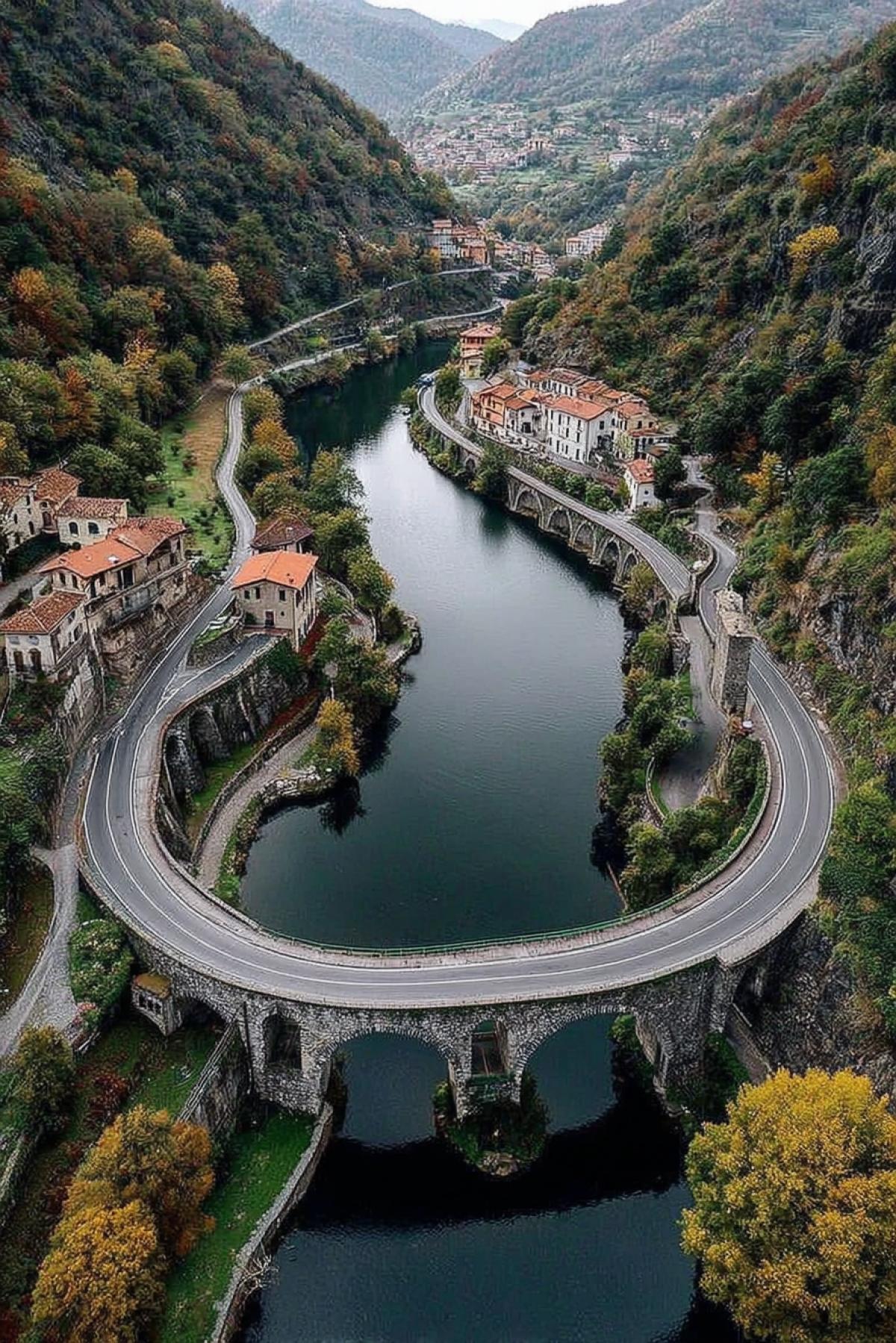 A picturesque aerial view of winding roads hugging the edge of a tranquil river, with a stone bridge arching over the water. The road is lined with colorful red-roofed houses, and the surrounding hilly landscape is lush with trees and autumn greenery, suggesting a tranquil countryside. The calm waters of the river reflect the clear sky above, and the arches of the bridge add a sense of architectural charm to the scene. A distant view of a small town at the foot of the hills.