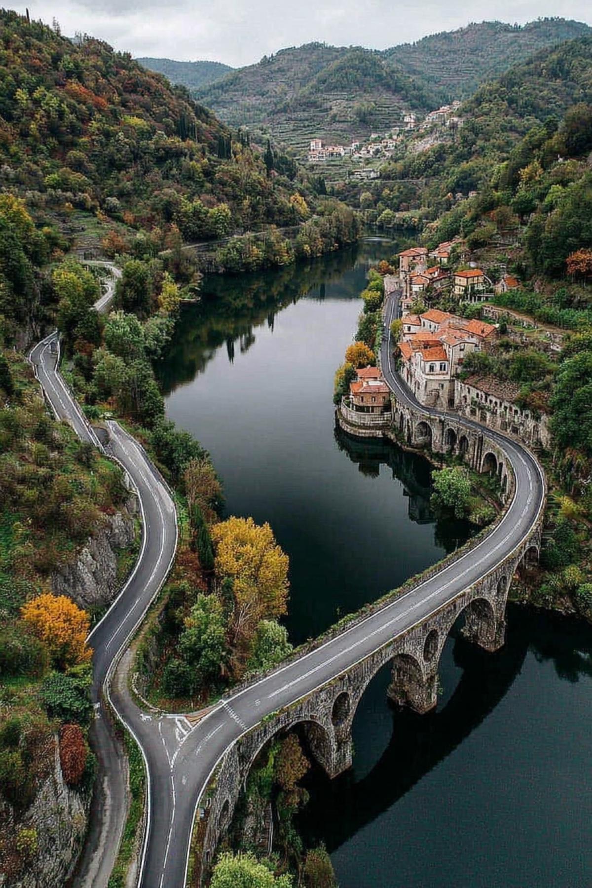 A picturesque aerial view of winding roads hugging the edge of a tranquil river, with a stone bridge arching over the water. The road is lined with colorful red-roofed houses, and the surrounding hilly landscape is lush with trees and autumn greenery, suggesting a tranquil countryside. The calm waters of the river reflect the clear sky above, and the arches of the bridge add a sense of architectural charm to the scene. A distant view of a small town at the foot of the hills.