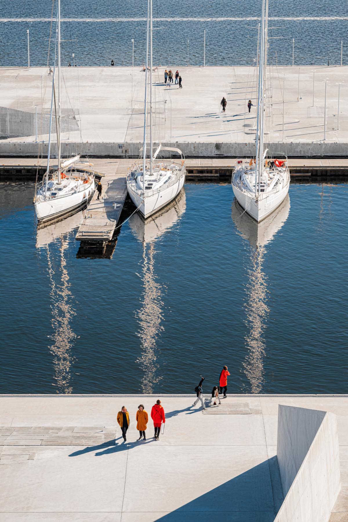 A number of snowwhite yachts at the modern concrete pier, their masts are reflected in smooth water. Geometric shadows from the railing, the glare of the sun on the waves. People in bright clothes (red, yellow) revive the scene. Filmed on a wide-angle, style: architectural photography with pop art accents.