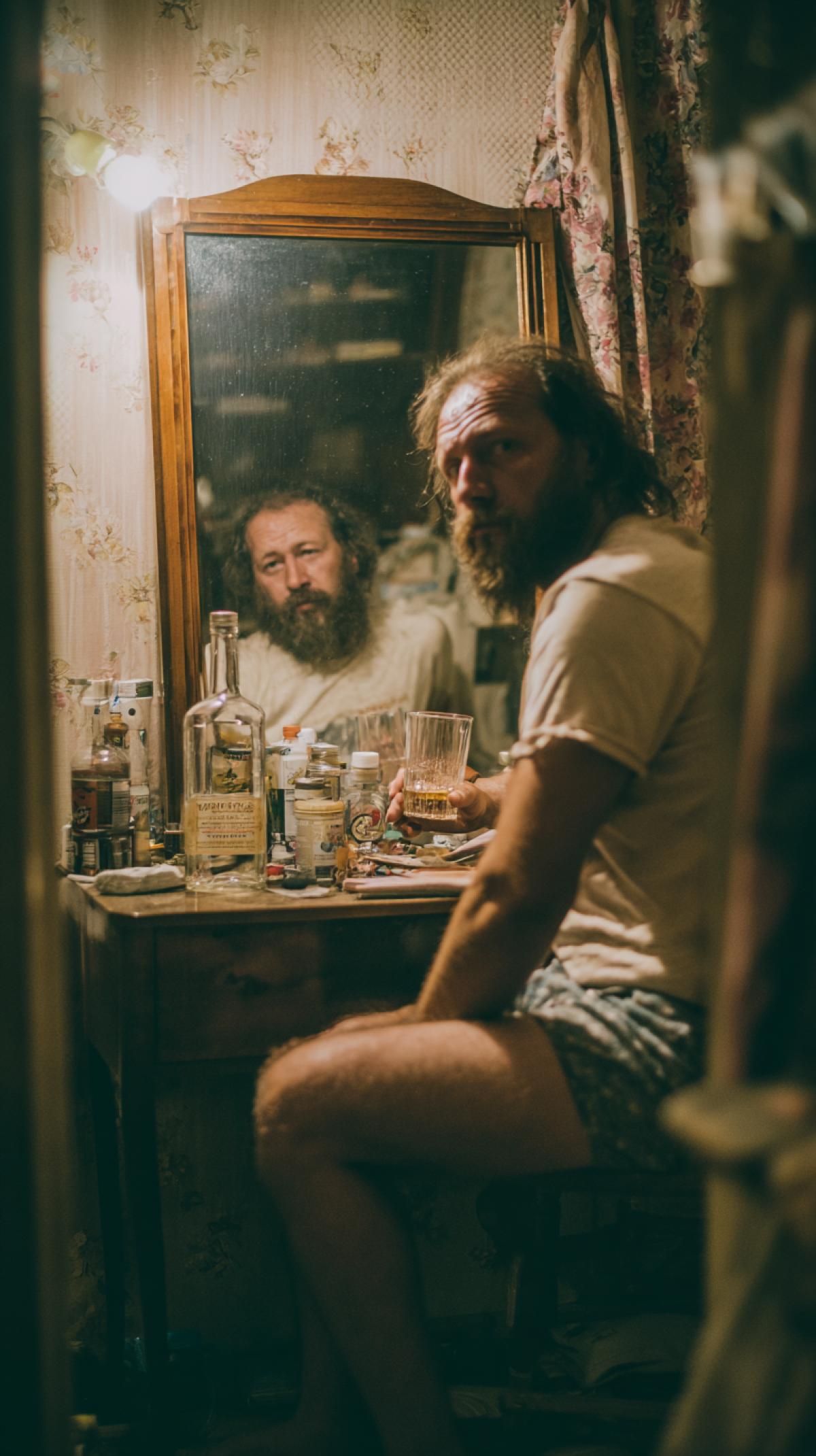 A man sits at the table, a drunk in a Tshirt and underpants, overgrown and dirty with a beard, a glass of vodka and a bottle of vodka, pronounces a toast, looking into his reflection in the mirror