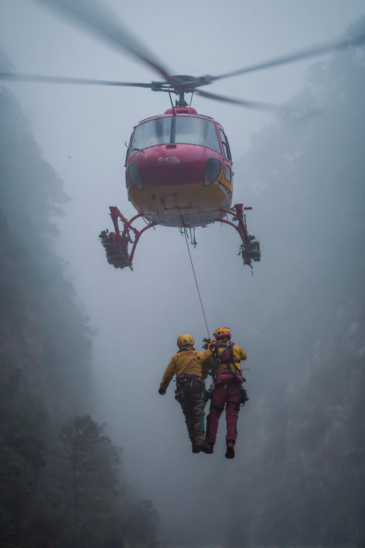 High-altitude medevac team suspended from a rope under a storm-chopper, lowering into a foggy mountain gorge where a wrecked cable car hangs over the abyss one rescuer clings to the cable, reaching for an injured climber, lightning flashes in clouds above, extreme vertical com4k, hyperreal, heart-stopping moment