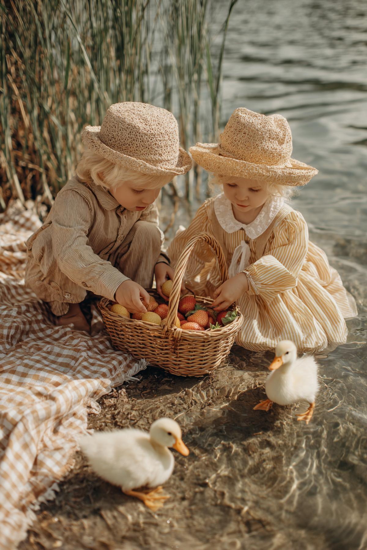 A happy family picnic on the shore of the lake: two small peanuts (a boy and a girl 3-4 years old) in straw panamas, they are bought with cookies on a checkered blanket while parents pour lemonade. One baby pulls a hand to a floating duckling, the other laughs, dropping the berry into a basket of strawberries. Warm sun bunnies play in water glare, the air is filled with the aroma of freshly cut grass. The style of the retro-opening of the 1950s with a soft watercolor texture, but in 8k quality. Touching scene of the perfect summer day