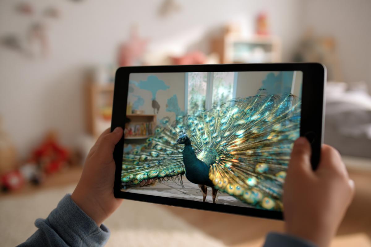 A close-up of a tablet held by a child’s hands, its screen displaying a vibrant, animated peacock with a shimmering tail bursting into a modern children’s room interior. The peacock appears lifelike with green, blue, and gold feathers, moving within the frame. The child’s hands grip the tablet, surrounded by the same room decorlight walls, toys, a bedbut without the peacock. The camera focuses on the screen, emphasizing the augmented reality effect. Soft daylight creates a modern, futuristic atmosphere with an unusual perspective.