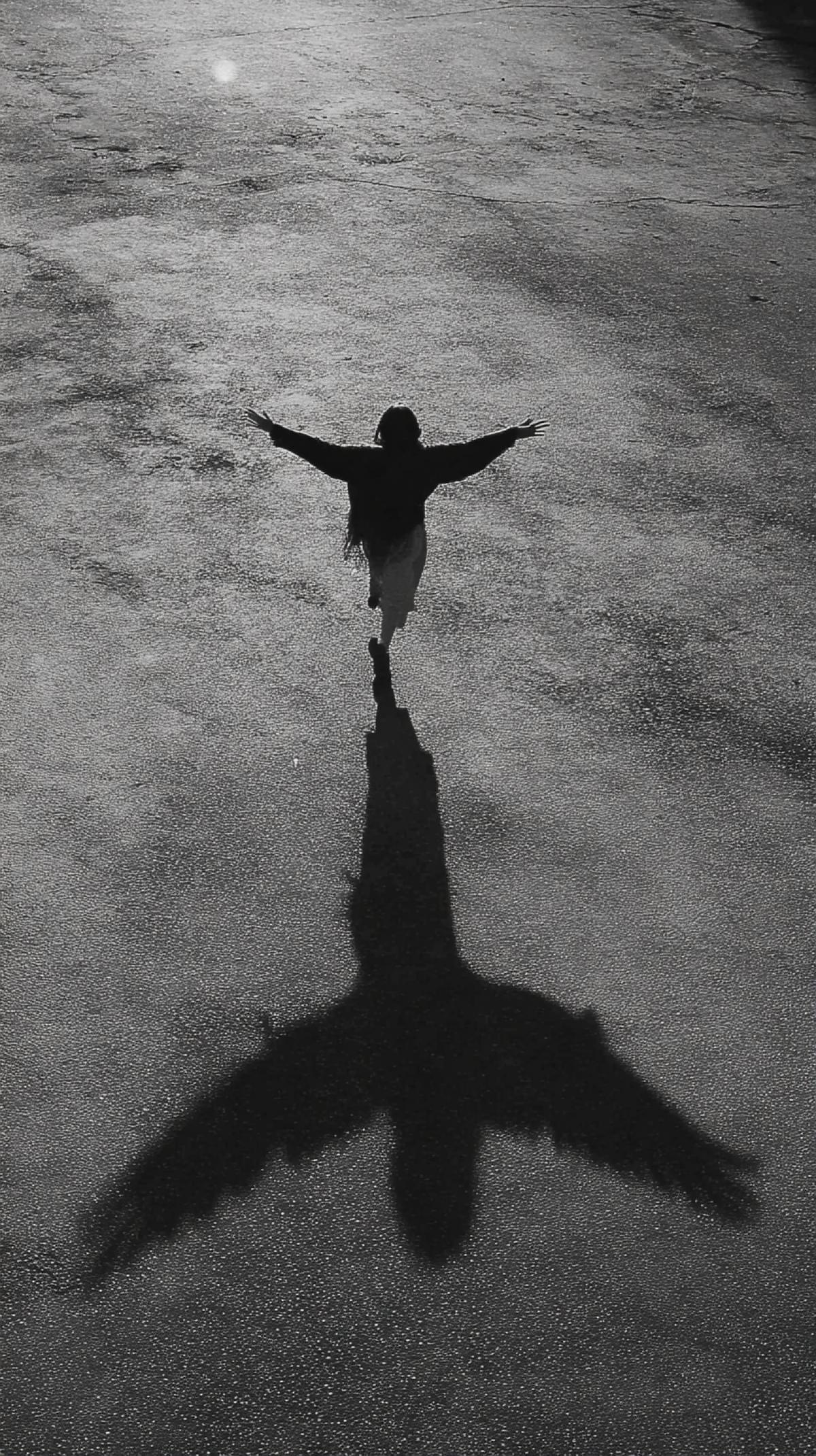 The girl runs with wideopen hands, her long shadow falls to the ground in the form of a silhouette of an eagle with open wings. dark and white photography. The photo is taken from above, the angle creates a feeling of depth and prospects. Minimalism, with a simple background, in the style of Daido Moriama, using the Leica M6 camera, f/8 diaphragm and the effect of granular film.