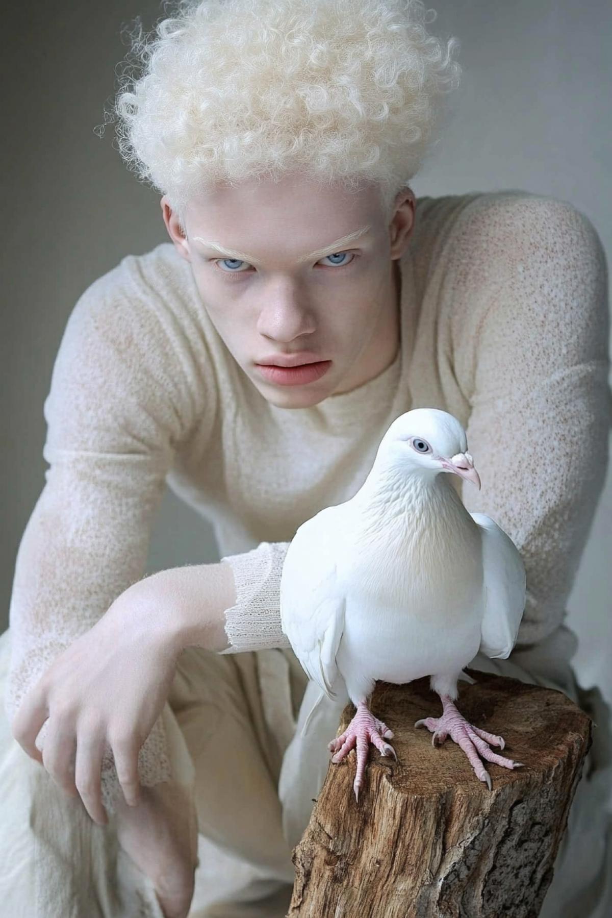 On a reclining piece of wood sits a pale young albino man with delicate freckles and white curly hair, blue transparent eyes, dressed in white clothes, on whose leg sits a large white dove.