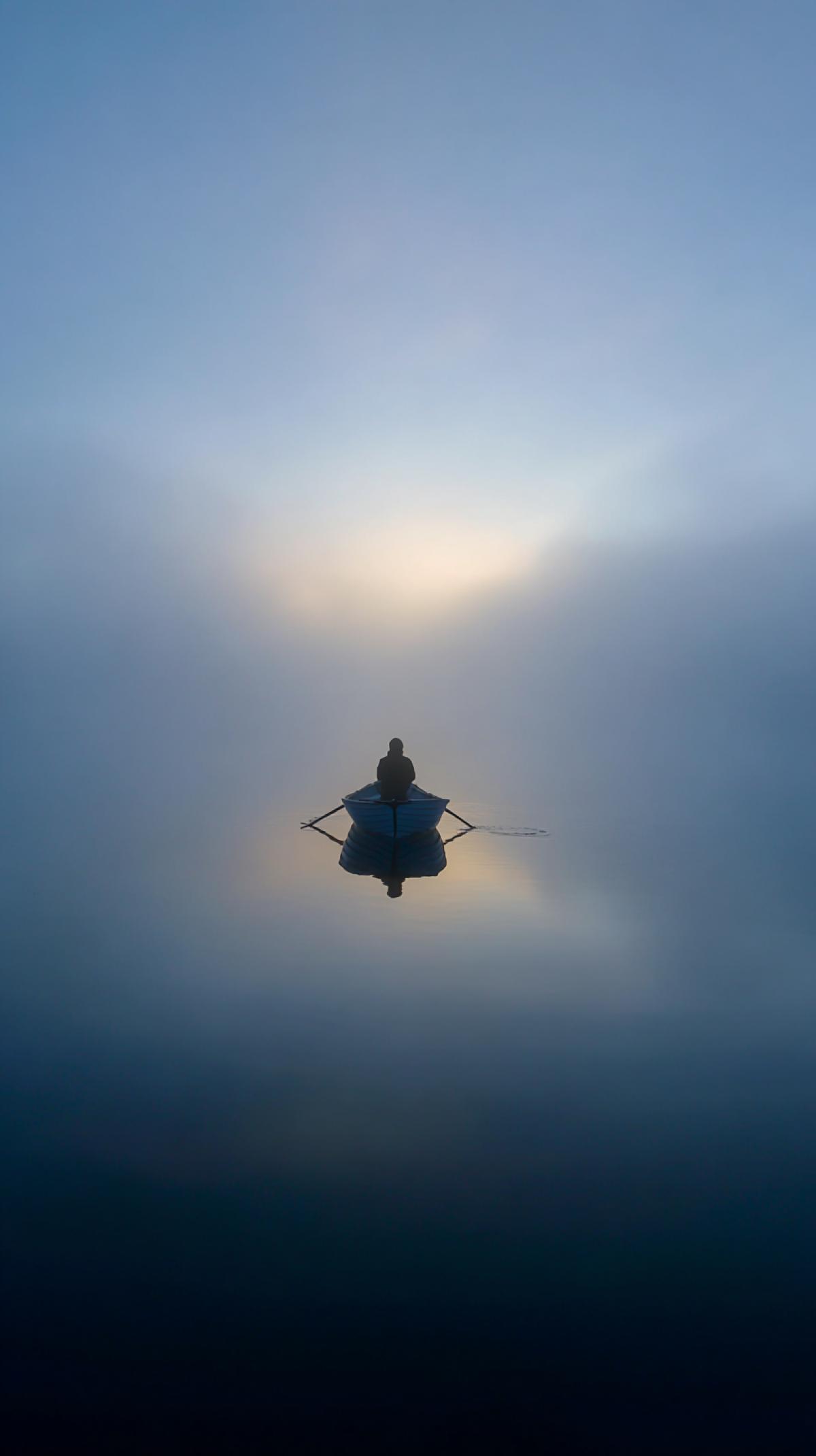 An atmospheric wide shot of an introverted man in a small boat, a solitary figure amidst a vast, calm lake. Its dawn, with a beautiful, thick fog creating a dreamlike, secluded sanctuary. The waters surface is perfectly still, showing a crisp, symmetrical reflection of the boat and the man. Soft, diffused morning light breaks through the mist, emphasizing the tranquility and peaceful escape. Serene, meditative, quiet, highly detailed