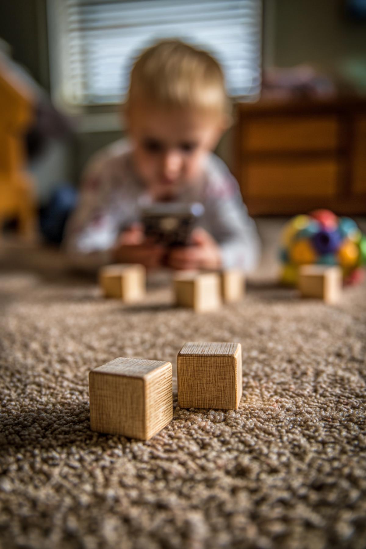 On the floor are childrens wooden plaything blocks in the foreground. In the background, a small child is blurryly looking at a smartphone.