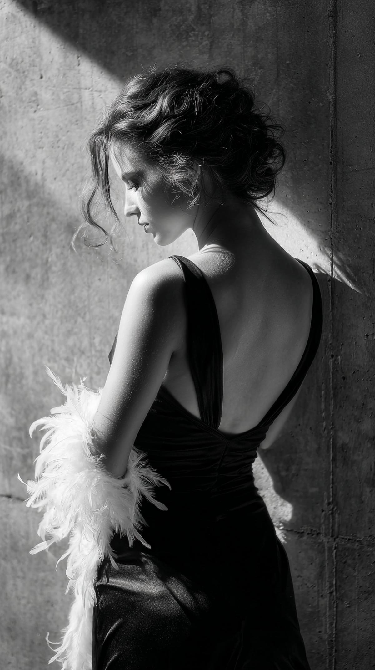 Scene Girl stands with her back to the camera against the background of a concrete wall, the head in the profile, the dress of the dress falls from the shoulder, the dark evening dress with the neckline on the back, the hair is collected in the bundle of the curls fall on the shoulders, white feathers, monochrome photos, realism, artistic image, play of shadows are flying