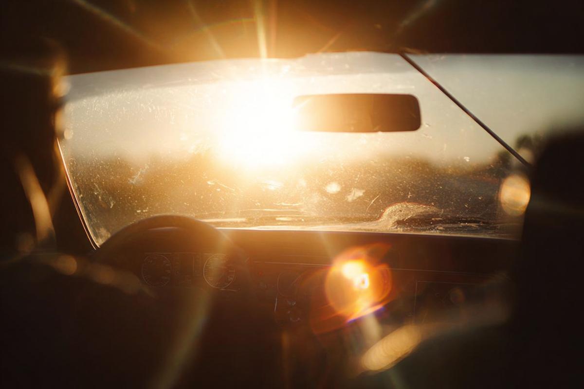 A breathtaking sunset viewed through a car windshield, the sun glaring intensely, creating lens flares and reflections on the glass. The interior of the car is silhouetted against the bright light. Photorealistic, dramatic lighting, 8k, warm colors, atmospheric, cinematic.