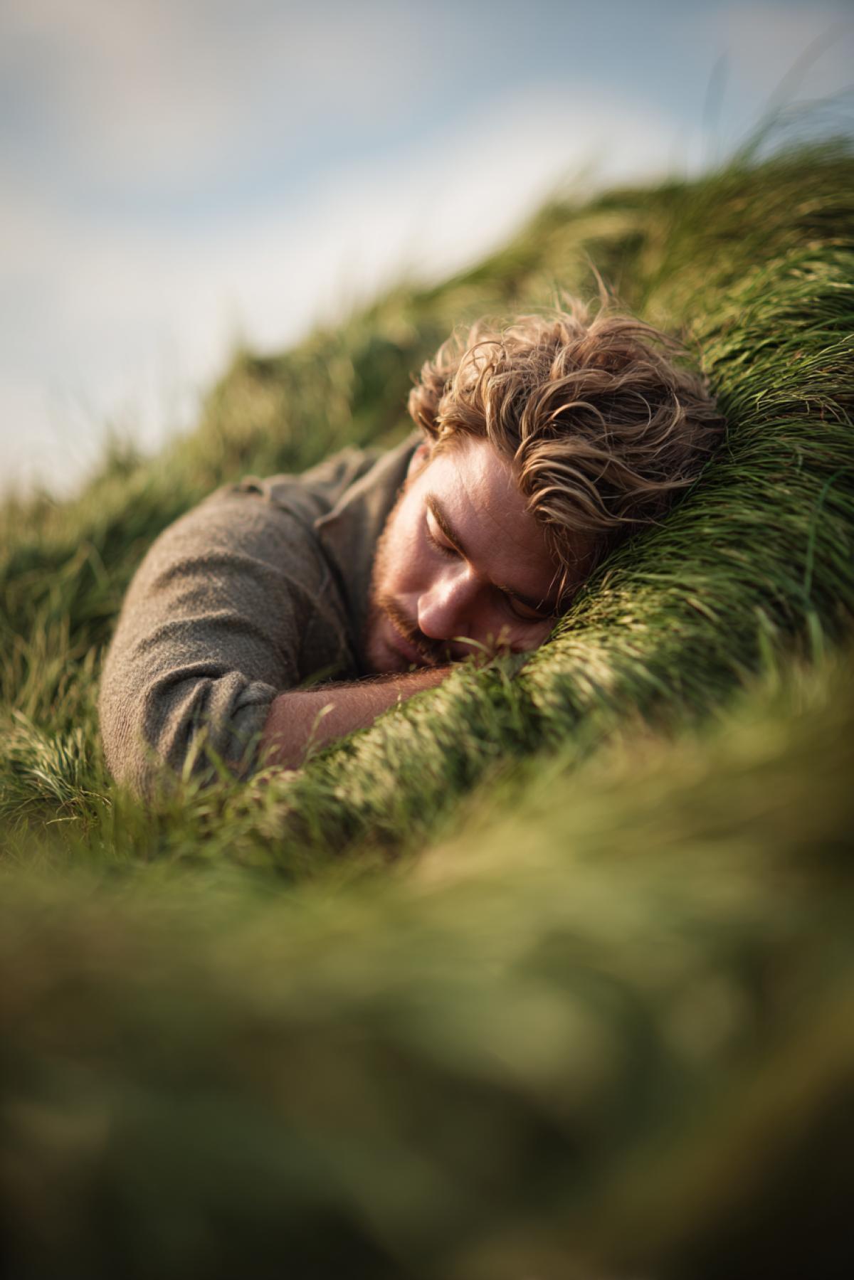 A man lying on his stomach, hugging a large green tussock (grass mound) in a meadow. His expression is peaceful and content, as if embracing the earth itself. Soft daylight, warm summer atmosphere, hyperrealistic, shallow depth of field, focus on the texture of the grass and the mans relaxed posture.