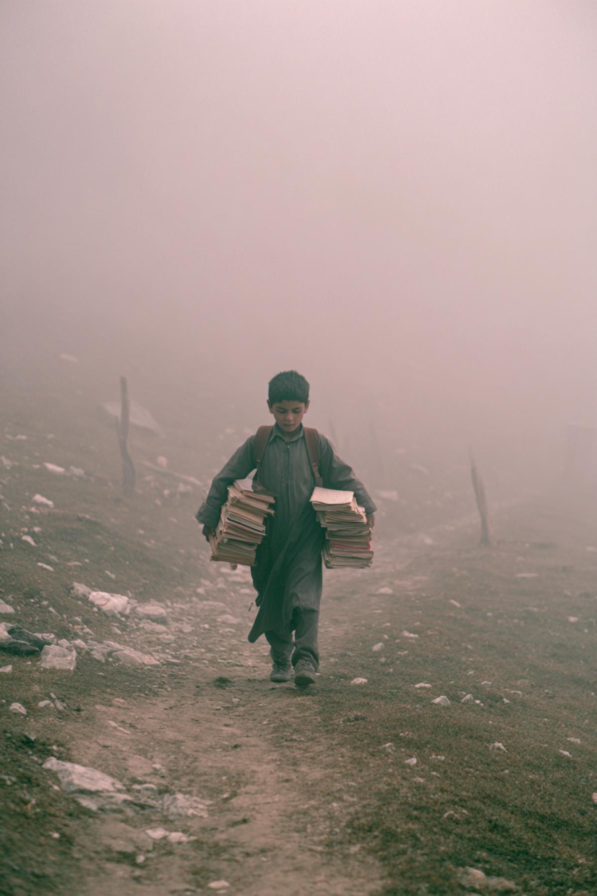 A young child walking a difficult path to school through a harsh mountainous landscape, carrying old books, determined expression, photorealistic, dramatic lighting, early morning fog, cinematic, shot on 35mm film, nostalgic atmosphere