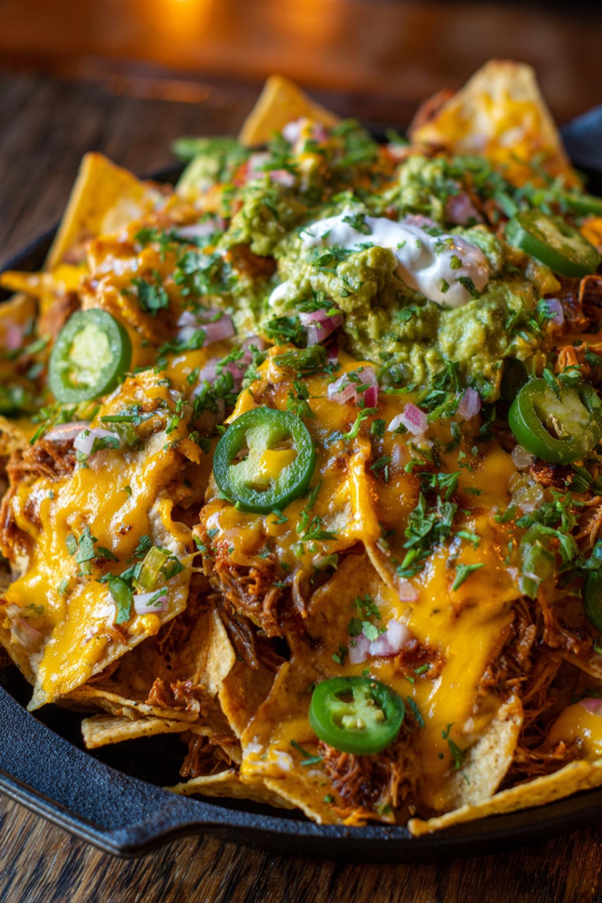 A chaotic and delicious pile of loaded nachos in a sports bar. A mountain of crispy tortilla chips overflowing with molten neon orange cheese, vibrant green jalapeños, creamy guacamole, tangy sour cream, and chunks of juicy pulled pork. Shot from above, top-down view, messy and inviting, shallow depth of field, warm ambient lighting, film grain, style of a vibrant food blog photo.
