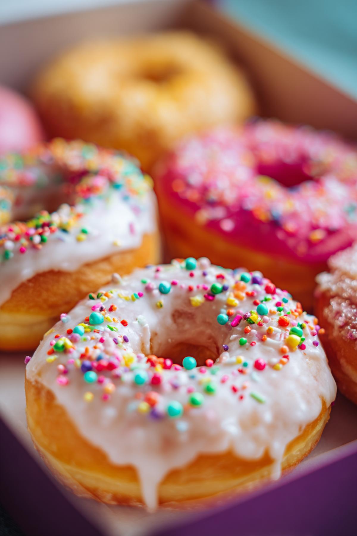 Box of perfect freshly glazed donuts, glossy sugar icing dripping slowly, variety of colorful sprinkles and rainbow sugar pearls, soft fluffy texture, cinematic lighting, shallow depth of field, vibrant colors, food photography style