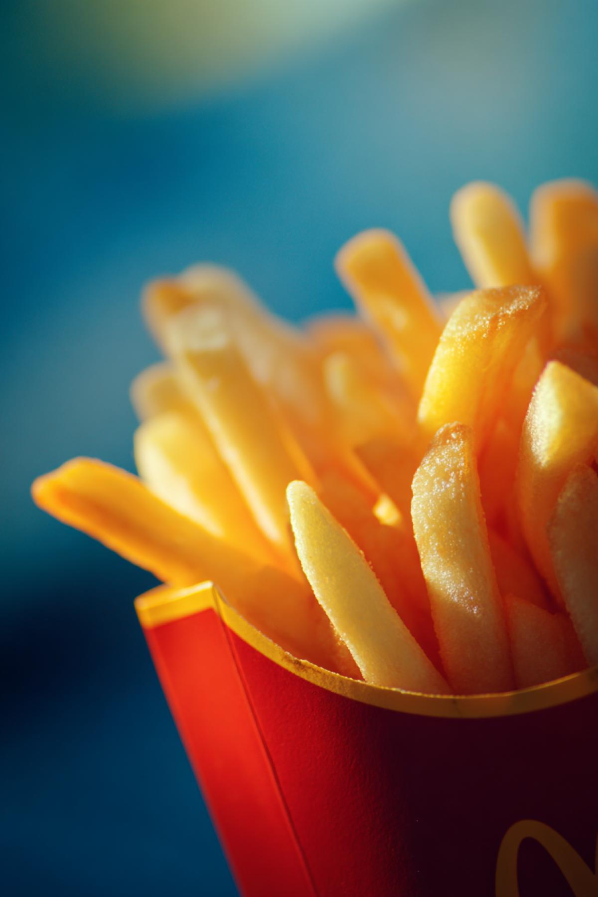 Fast food photo: bright packaging of McDonalds French fries. with shades of orange and. yellow. Light is focused around the packaging. The background is blue and slightly blurred