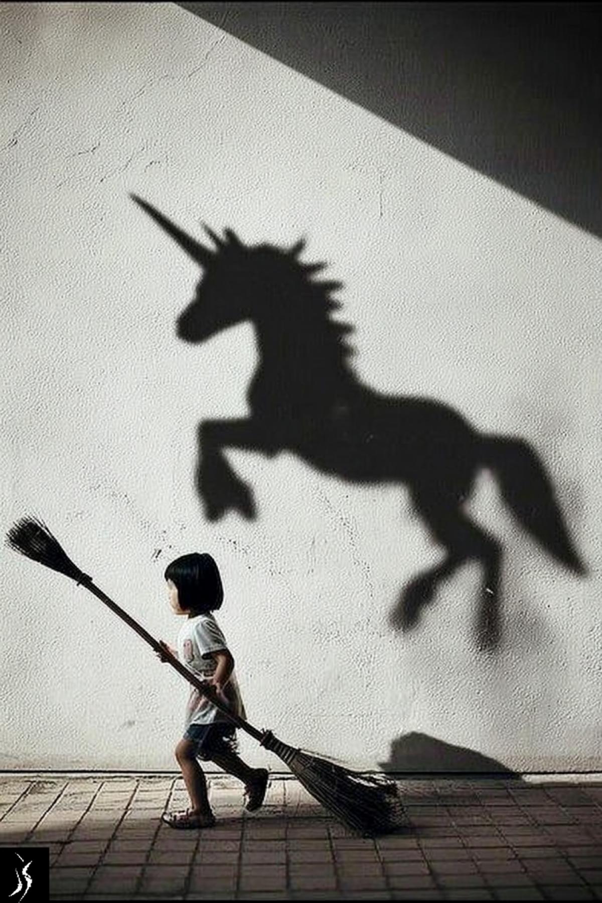 A little girl riding a broomstick, her shadow falls on the wall in the shape of a unicorn. dark and white photo. The photo was taken from the side, the angle creates a sense of depth and perspective. Minimalism, with a simple background, in the style of Daido Moriyama, using a Leica M6 camera, F/8 aperture, and a grainy film effect.