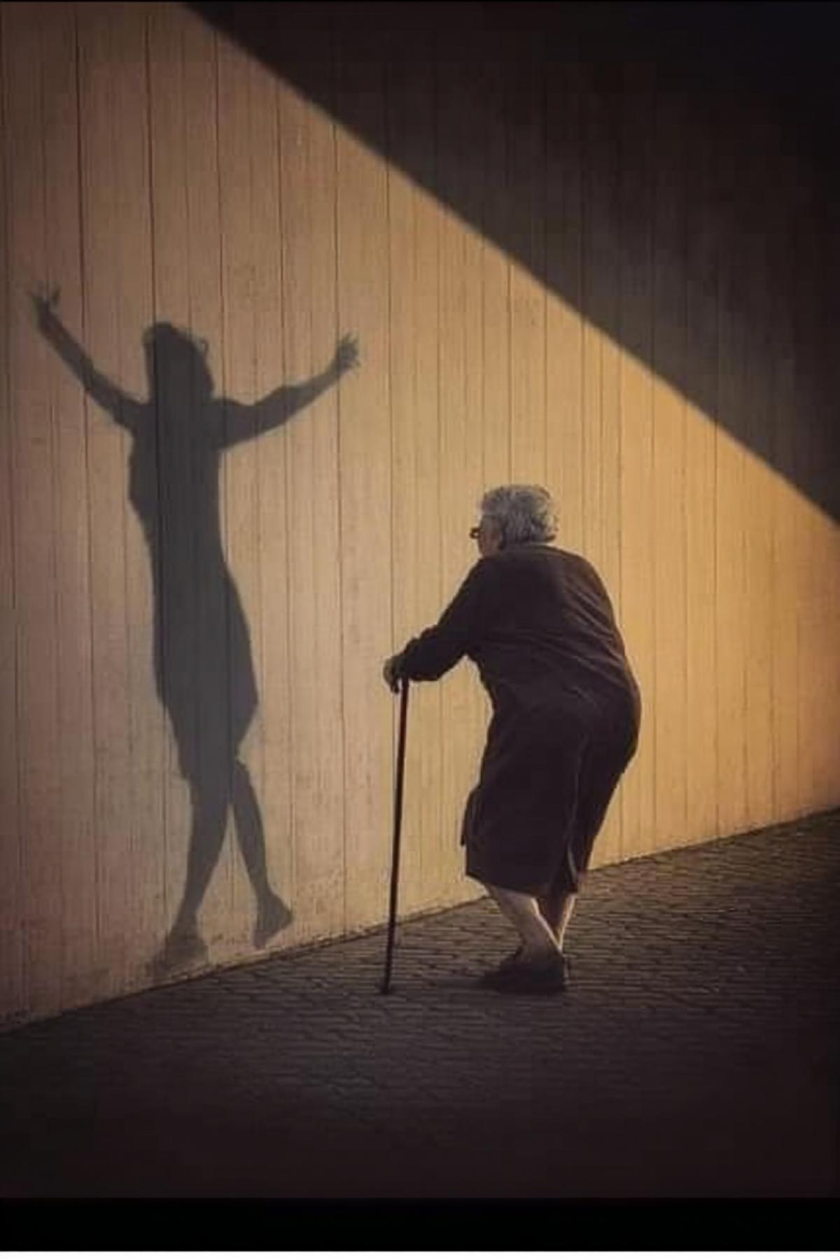 An old woman walks leaning on a stick, her shadow falls on the wall in the form of a silhouette of a young girl with her arms open. sepia photography. The photo was taken from the side, the angle creates a sense of depth and perspective. Minimalism, with a simple background, in the style of Daido Moriyama, using a Leica M6 camera, F/8 aperture and a grainy film effect.