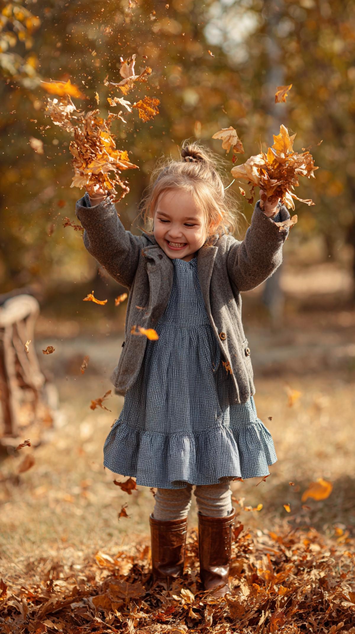 a little girl in a coat and dress, brown boots, having fun throwing autumn leaves over herself, photo, autumn colors, sunny day, park