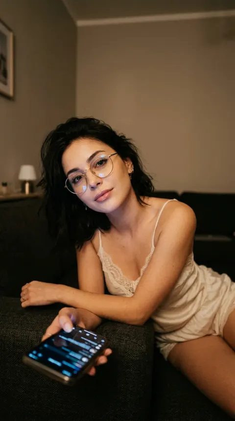 A young adult woman with soft refined features, thin metal glasses, and shoulder-length dark tousled hair, leaning forw…