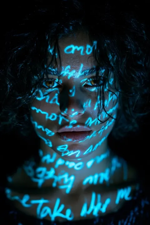 woman. a tight vertical portrait of a person with dark curly hair and hazel eyes, their face and collarbone illuminated…